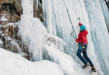 Ice climber dressed in warm winter climbing clothes, safety harness and helmet climbing frozen waterfall using two Ice climbing axes and crampons. Active people and sports activities concept image.の写真素材