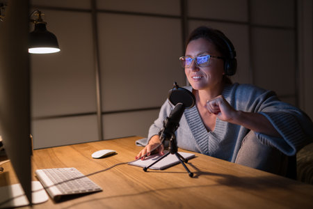 A middle-aged woman in headphones smiling and talking with somebody chatting video call using microphone with pop filter and desktop computer. Internet online video conference technology concept imageの写真素材