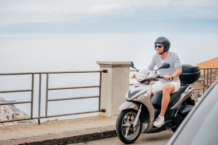 Cheerfully smiling man in helmet and sunglasses fast riding the moto scooter on the Sicilian old...