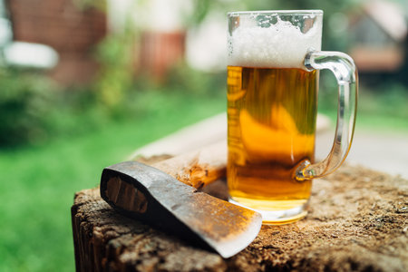 Still lifephoto of glass of fresh cold beer on wooden stub alongside axe. Rural concept, masculinity, coarse, rough wilderness lifestyle concept.の写真素材