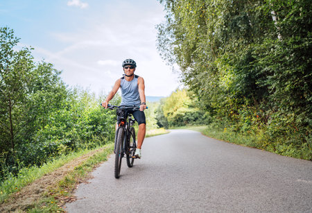 Portrait of a happy smiling man dressed in cycling clothes, helmet and sunglasses riding a bicycle on the asphalt out-of-town bicycle path. Active sporty people concept image.の写真素材