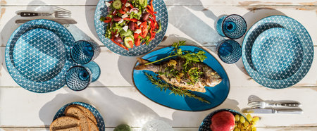 Gilt-head bream also known as Orata baked in the oven and served on a white wooden table with vegetable salad and fruits. Healthy seafood preparation and serving concept image.の写真素材