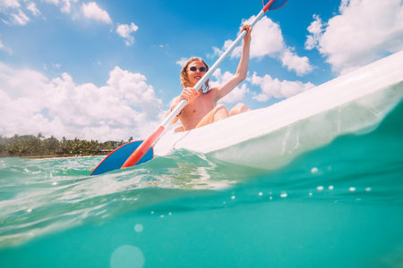 Underwater view angle to a smiling blonde teenage boy in fashion sunglasses rowing single sit kayak with palm grove background. Active people summer vacation sea concept image, Le Morne, Mauritius.の写真素材