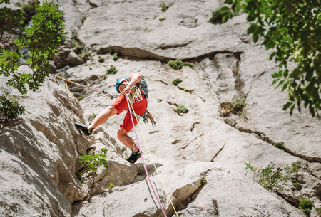 Active climber middle age man in protective helmet while abseiling from cliff rock wall using ropes with a belay device and climbing harness. Active extreme sports time spending conceptの写真素材