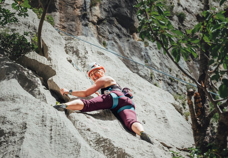 Smiling athletic woman in protective helmet climbing cliff rock wall using top rope and climbing harness in Paklenica National park site in Croatia. Active extreme sports time spending concept.の写真素材