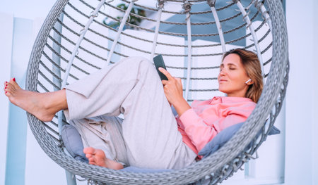 Beautiful middle-aged woman listening Earbuds smartphone music lying in comfortable hanging chair on open house terrace and enjoying the sunset sun rays. Modern technology and home lifestyle conceptの写真素材