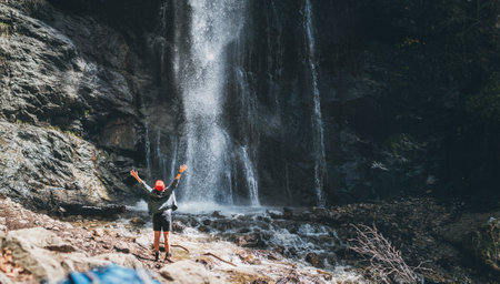 A woman dressed in active trekking clothes standing near mountain river waterfall rose arms up and enjoying the splashing Nature power. Backpack on foreground. Traveling, trekking nature concept imageの写真素材