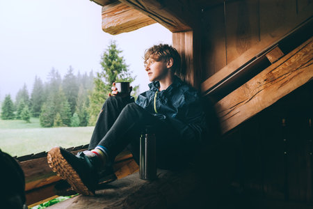 Young lonely teenager boy drinking hot tea from thermo flask while he sitting on the wooden forest house terrace and enjoying the morning spruce foggy forest. Traveling and tourism at fall time.の写真素材