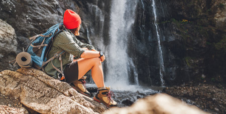 Woman with a backpack in a red hat dressed in active trekking clothes sitting near the mountain river waterfall and enjoying the splashing Nature power. Traveling, trekking, and a nature concept imageの写真素材