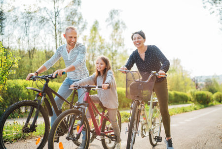 Smiling father and mother with daughter during summer outdoor bicycle riding. They enjoy togetherness in the summer city park. Happy parenthood and childhood or active sport life concept image.の写真素材