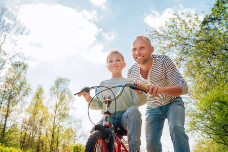 Smiling father teaching his daughter to ride a bicycle. They enjoy togetherness in the summer city park. Happy childhood concept image.の写真素材