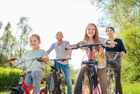 Smiling father and mother with two daughters during summer outdoor bicycle riding. They enjoy togetherness in the summer city park. Happy parenthood and childhood or active sport life concept image.の写真素材