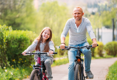 Portraits Smiling father with daughter during summer outdoor bicycle riding. They enjoy togetherness in the summer city park. Happy parenthood and childhood or active sport life concept image.の写真素材