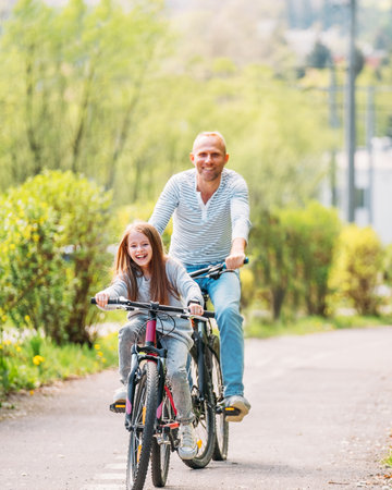 Portraits Smiling father with daughter during summer outdoor bicycle riding. They enjoy togetherness in the summer city park. Happy parenthood and childhood or active sport life concept image.の写真素材