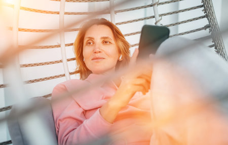Beautiful middle-aged woman portrait listening earbuds using modern smartphone music sitting in a comfortable hanging chair. Modern technology and home lifestyle conceptの写真素材