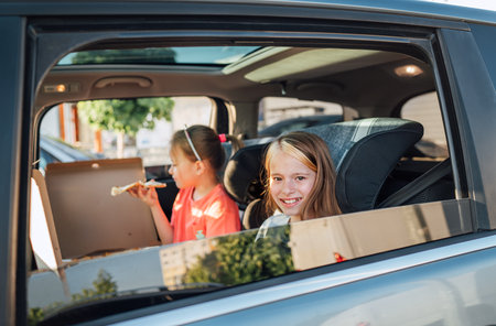 Two positive smiling sisters are happy to eat just cooked Italian pizza sitting in child car seats on the car back seat. Happy childhood, fast food eating, or auto journey lunch break concept image.の写真素材