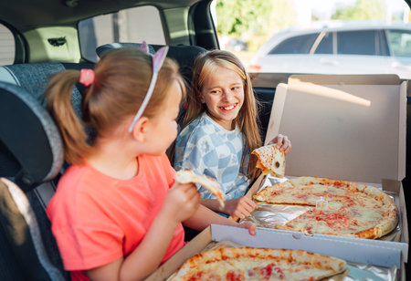 Portrait of two positive smiling sisters eating just cooked italian pizza sitting in child car seats on car back seat. Happy childhood, fastfood eating or auto jorney lunch break concept image.の写真素材