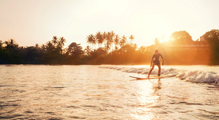 Teen boy silhouette riding a long surfboard. He caught a wave in an Indian ocean bay with magic sunset background. Extreme water sports and exotic countries concept.の写真素材