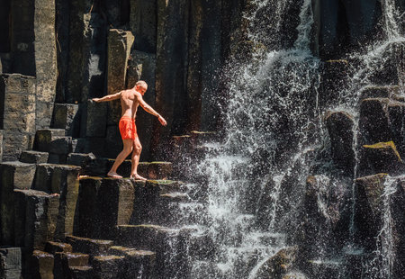 Caucasian man in swimsuit balancing near the falling water streams flowing on black volcanic stone cascades. Rochester Falls waterfall - popular tourist spot in Savanne district in Mauritius.の写真素材