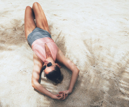 Sunbathing middle-aged woman in a swimsuit and fancy sunglasses lying in palm tree shadows on the sandy ocean beach. Healthy tanning and an exotic vacation concept image.の写真素材