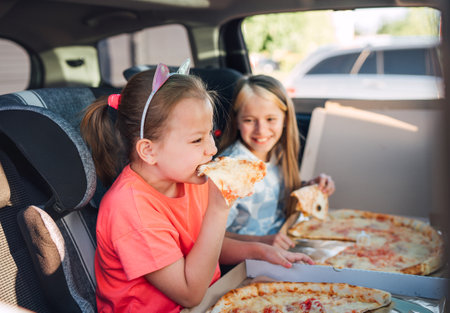 Portrait of two positive smiling sisters eating just cooked italian pizza sitting in child car seats on car back seat. Happy childhood, fastfood eating or auto jorney lunch break concept image.の写真素材
