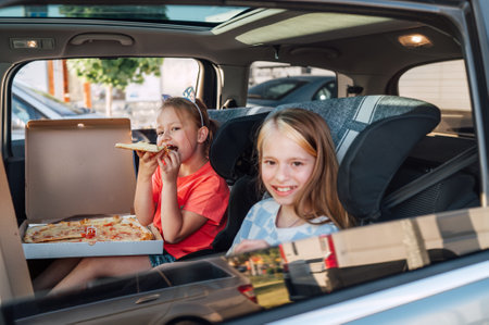 Two positive smiling sisters are happy to eat just cooked Italian pizza sitting in child car seats on the car back seat. Happy childhood, fast food eating, or auto journey lunch break concept image.の写真素材
