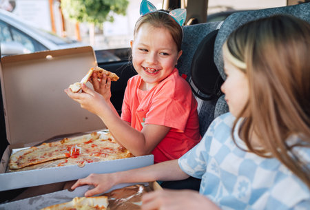 Portrait of two positive smiling sisters eating just cooked Italian pizza sitting in child car seats on the car back seat. Happy childhood, fast food eating, or auto journey lunch break concept image.の写真素材