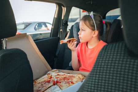 Portrait of little 5 YO girl eating just cooked Italian pizza sitting in child car seat on car back seat and looking out window . Happy childhood, fast food eating, or auto journey lunch break imageの写真素材