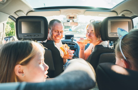 Happy smiling young couple with two daughters eating just cooked Italian pizza sitting in modern car. Happy family moments, childhood, fast food eating or auto journey lunch break concept image.の写真素材