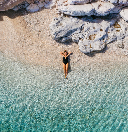 Top view photo showing a woman dressed black swimsuit lying on a white pebble lonely beach, calm waves washing up her legs. Exotic tropical summer holidays concept. Cephalonia island, Greece.の写真素材