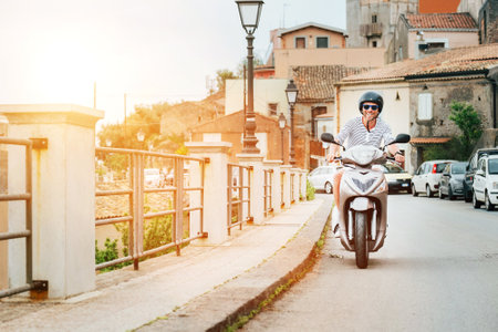 Cheerfully smiling man in helmet and sunglasses fast riding the moto scooter on the Sicilian old town streets in the Forza d'Agro. Happy Italian vacation and transportation concept.の写真素材