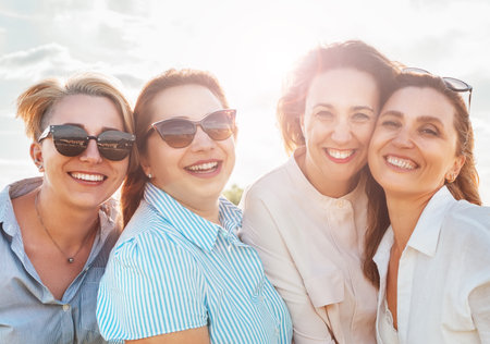 Half-length portraits of four cheerful smiling women in sunglasses embracing and looking at camera during outdoor walking with bright sun. Woman's friendship, relations, and happiness concept imageの写真素材