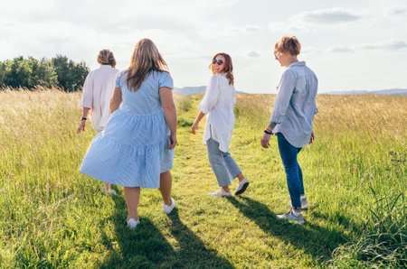 Four cheerful smiling women walking by the high green grass meadow hill, laughing and chatting with each other during a sunset walk. Woman's friendship, relations, and happiness concept image.の写真素材