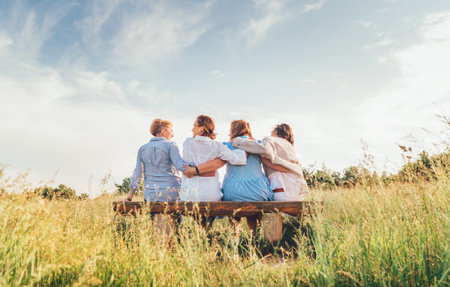 Four smiling women dressed light summer clothes harm embracing sitting on the meadow bench during outdoor walking. Woman's friendship, relations, and happiness concept image.の写真素材