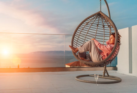 Beautiful middle-aged barefoot woman lying in comfortable hanging chair on open house terrace and enjoying the sunset sun rays. Modern home lifestyle and mental health concept imageの写真素材