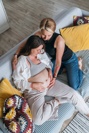 Young woman tender touching partner's female pregnant belly. Same-sex marriage couple in the home living room.  Woman's health, happy pregnancy doula supporting and calm mental mood concept image.の写真素材