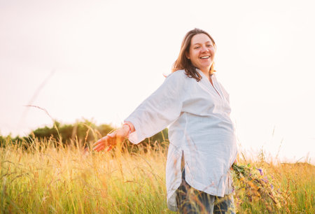 Sincerely smiling young pregnant woman dressed in light summer clothes walking by the high green grass meadow with wildflowers bouquet in evening sunset hours. Human in the nature concept image.の写真素材