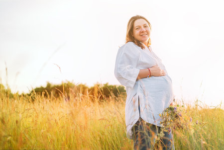 Sincerely smiling young pregnant woman dressed in light summer clothes walking by the high green grass meadow with wildflowers bouquet in evening sunset hours. Human in the nature concept image.の写真素材