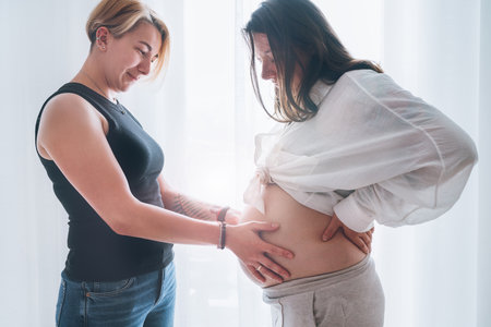 Young woman tender touching partner's female pregnant belly. Same-sex marriage couple next to living room window. Woman's health, happy pregnancy doula supporting and calm mental mood concept image.の写真素材