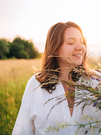 Sincerely smiling young pregnant woman dressed in light summer clothes walking by the high green grass meadow with wildflowers bouquet in evening sunset hours. Human in the nature concept image.の写真素材