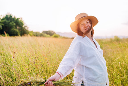 Sincerely smiling young pregnant woman dressed in light summer clothes and straw hat walking by high green grass meadow with wildflowers bouquet in evening sunset hours. Human in nature concept image.の写真素材