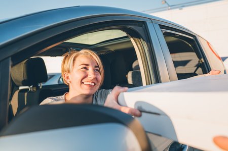 Young female sitting in car driver seat and sincerely smiling to deliveryman accepting two pizza boxes on the city street. Small business, people relations, fast foot meal consumption concept photoの写真素材