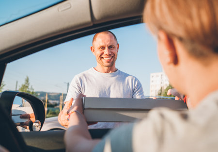 Young sincerely smiling deliveryman passing two pizza boxes to female sitting in car driver seat to on the city street. Small business, people relations, fast foot meal consumption concept photoの写真素材