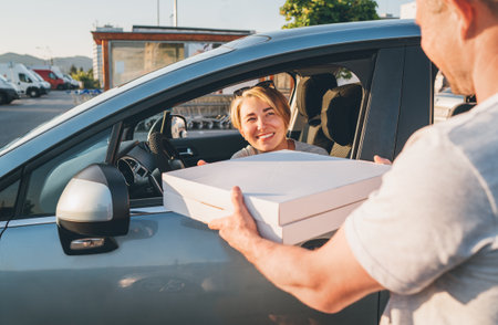 Young female sitting in the car driver seat and smiling to deliveryman accepting two pizza boxes on the city street. Small business, people relations, fast foot meal consumption concept photo.の写真素材