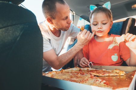 Father wiping gently daughter's mouth while eating just cooked Italian pizza during family car trip brake stop. Happy family moments, childhood, fast food eating or auto journey lunch break conの写真素材
