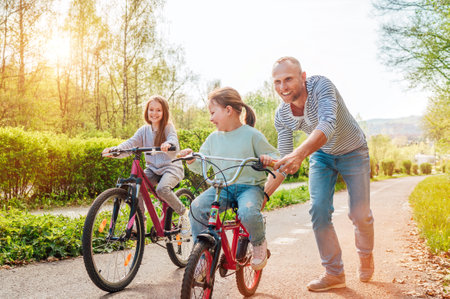 Smiling father with two daughters during outdoor walk. He teaching younger girl to ride a bicycle. They enjoy togetherness in the summer city park. Happy childhood concept image.の写真素材