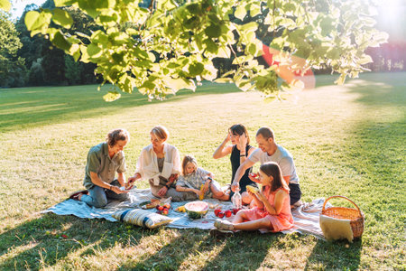 Big family under Linden tree on the picnic blanket on the in city park green grass. They are eating boiled corn, apples, peaches, pastries and watermelon. Family values and outdoor activities concept.の写真素材