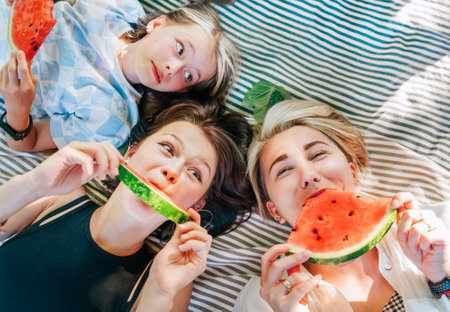 Mother lying with daughters lying on picnic blanket in city park during weekend sunny day, smiling, laughing at camera and eating juicy watermelon. Family values, fruits vitamins, outdoor time conceptの写真素材