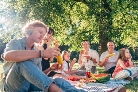 Big family sitting on the picnic blanket in city park during weekend Sunday sunny day. They are smiling, laughing and eating boiled corn and watermelon. Family values and outdoors activities concept.の写真素材