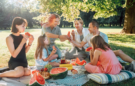 Big family sitting on the picnic blanket in a city park during weekend Sunday sunny day. They are smiling, chatting, laughing and eating juicy watermelon. Family values and outdoor activities concept.の写真素材